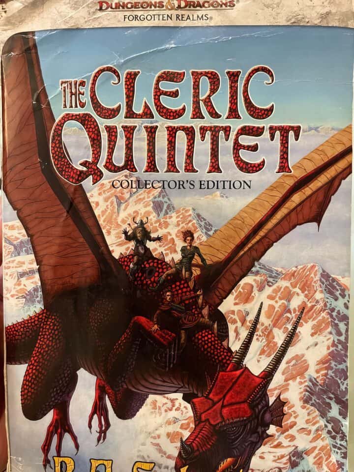 A young man is reading old books at a wooden table in a cozy study. The Cleric Quintet by R. A. Salvatore