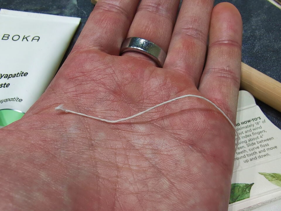 RADIUS Silk Floss Review: 5 Pros, 3 Cons & Eco-Friendly Truth 3 A hand with a silver ring shows a loose dental floss on the palm, with a tube of natural toothpaste and a wooden stick in the background, symbolizing oral hygiene and dental care.