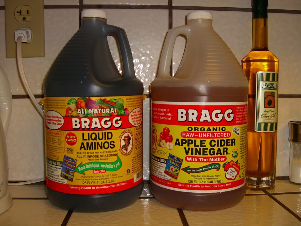 Liquid amino acids and organic apple cider vinegar bottles on a kitchen counter, used for cooking and health benefits, with olive oil and other condiments in the background.