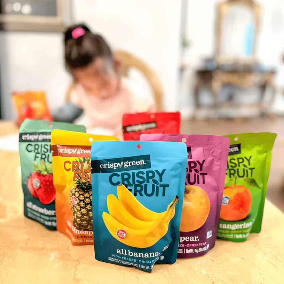 Crispy Green Healthy Habits Monthly Supply Box Review: How Freeze-Dried Fruit Won Over My Picky Eaters 2 Brightly colored Crispy Green fruit snack pouches on a table with a blurred background of a young girl.