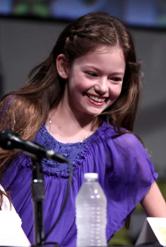 Easy-to-read alt text: Smiling young girl in purple blouse at a public event, with a water bottle and microphone nearby, showcasing happiness and engagement.