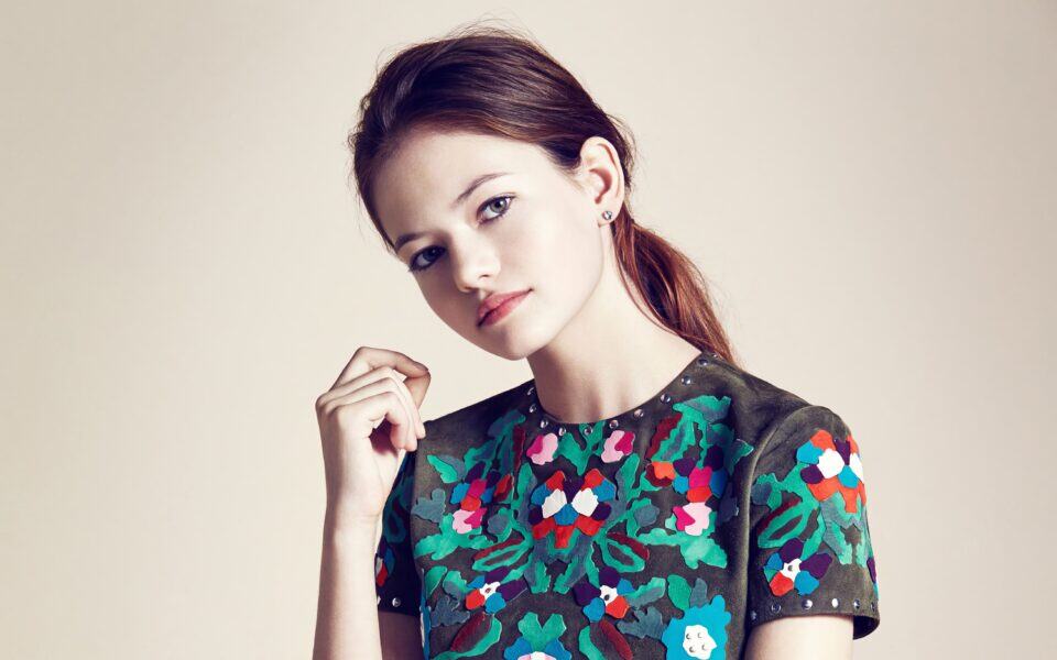 A young woman with red hair and fair skin poses in a stylish, embroidered floral top against a neutral background. Her confident expression and subtle makeup highlight modern fashion trends and elegant style.