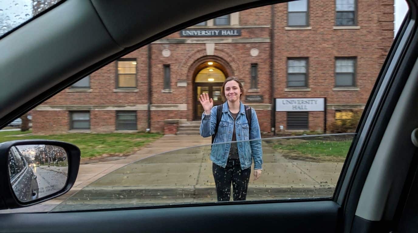View from inside a car of a college student waving goodbye in front of a dorm building. Young woman waving at camera outside university hall, student life, college campus, university experience, education journey, student portrait, traveling student, university building, campus visit, expanding knowledge.