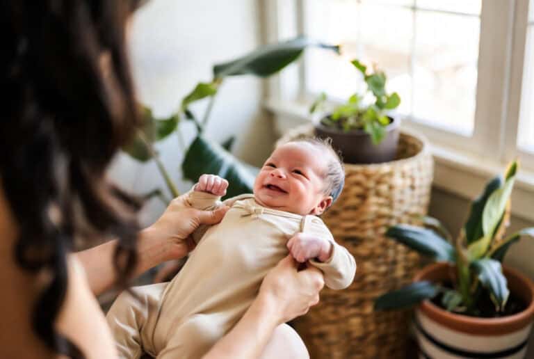 Close-up of a smiling baby being held by a caregiver at home. The baby is happy and relaxed, surrounded by lush houseplants near a bright window, capturing family moments.