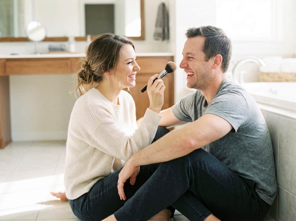 The Happy Wife's Guide to Sissy Husband Lifestyles 8 A happy couple enjoying a lighthearted moment at home, with the woman playfully holding a makeup brush near the man's face, symbolizing trust and connection in their relationship.