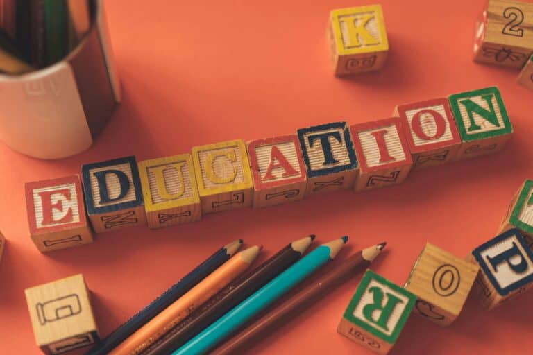 Colorful wooden blocks spelling "EDUCATION" on a pink surface with pencils nearby, symbolizing learning and challenging traditional education methods.