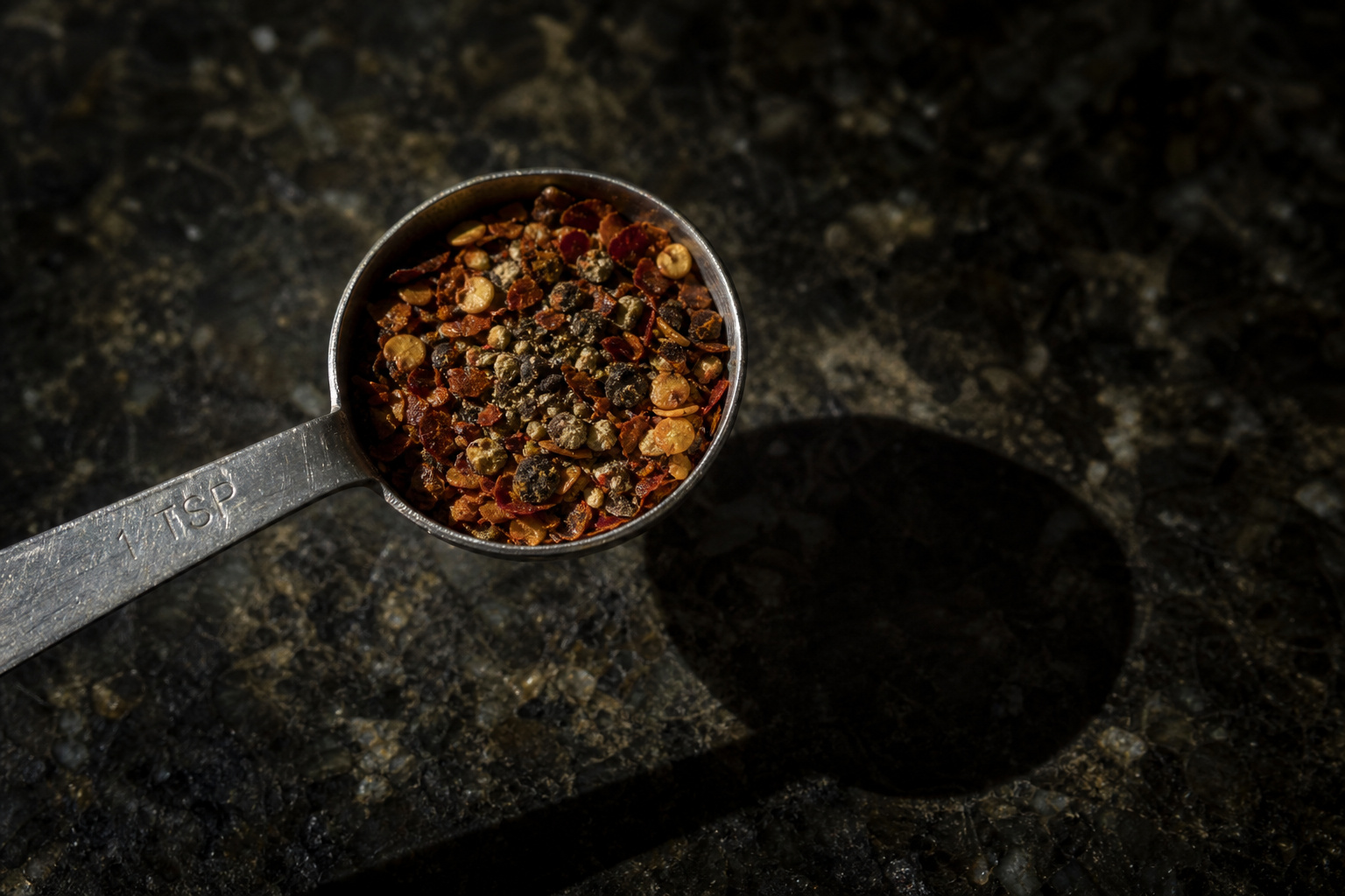 Close-up of a metal measuring spoon filled with mixed spices on a dark textured surface, casting a shadow.