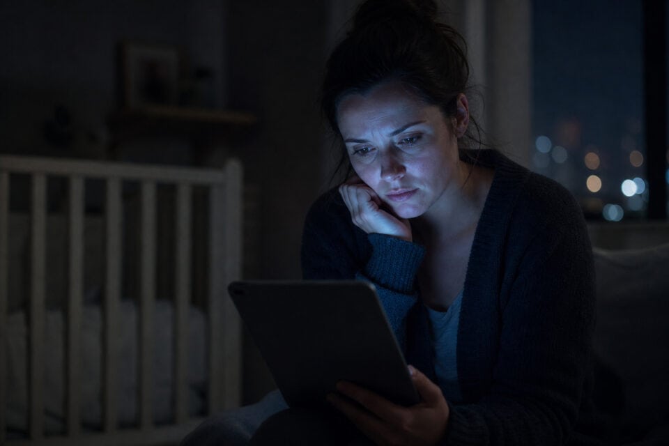 How to Be a Good Mom by Hitting the 70% Rule, Not 100% 2 Woman sitting in a dimly lit room at night, focused on her tablet, with a crib in the background, illustrating late-night parenting or relaxation.