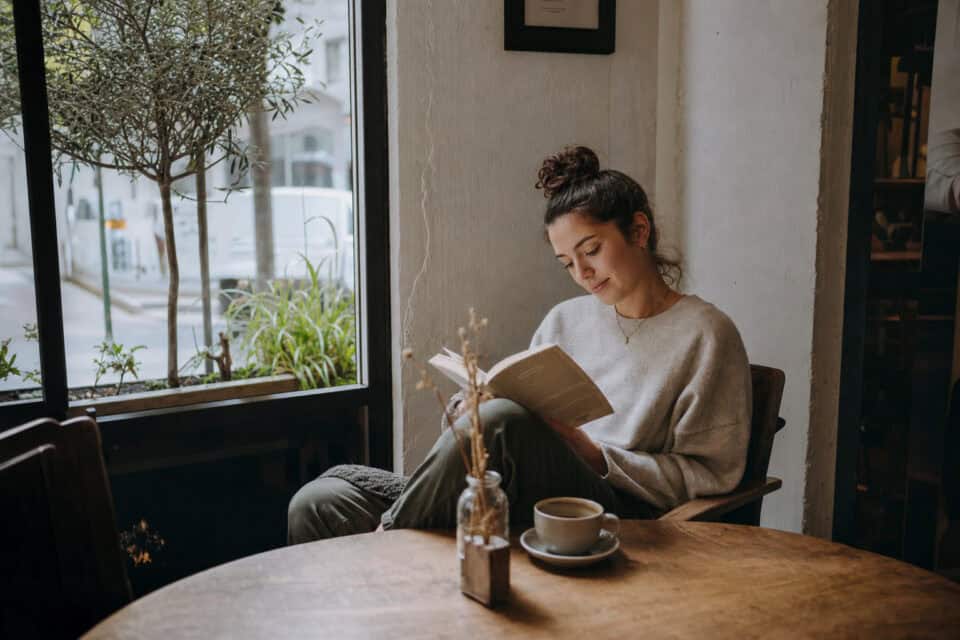 How to Be a Good Mom by Hitting the 70% Rule, Not 100% 9 Woman enjoying a peaceful reading session in a cozy café, sitting by a large window with natural light, surrounded by plants and warm decor.