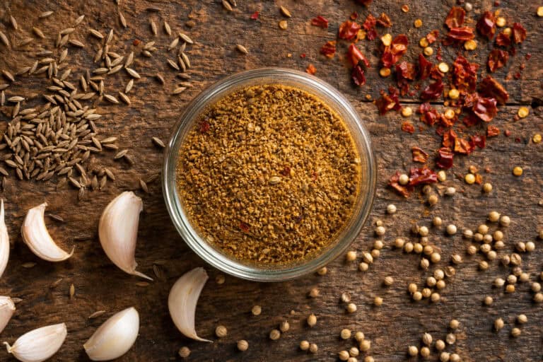 A glass bowl of ground spice surrounded by garlic cloves, cumin seeds, dried chili flakes, and white peppercorns on a rustic wooden background.