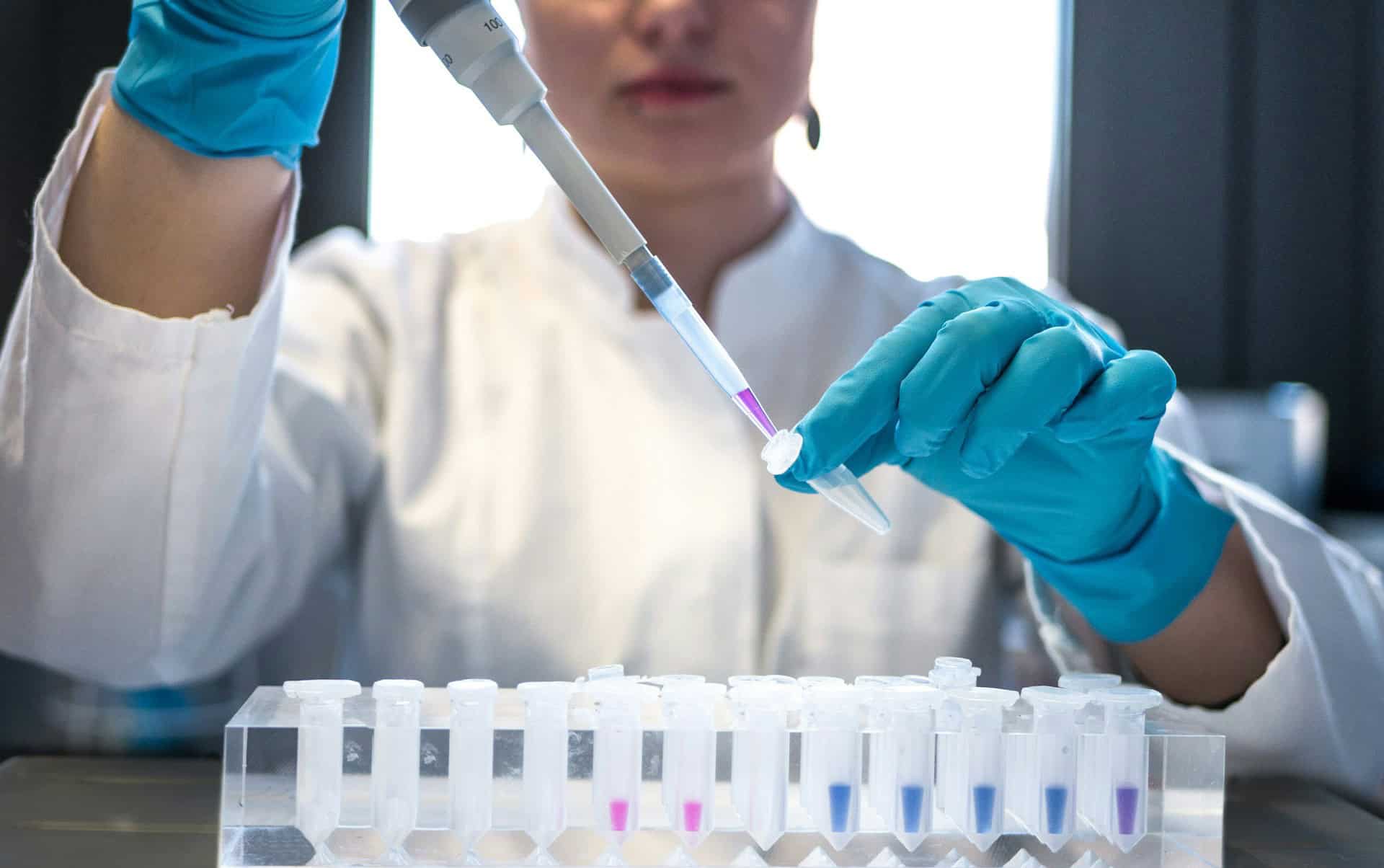 Laboratory scientist wearing gloves using a pipette to transfer liquid into test tubes in a scientific lab setting.