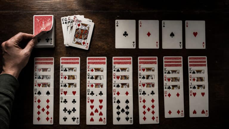 Close-up of a hand holding a playing card, with a spread of playing cards on a dark wooden table, including aces and numbered cards.