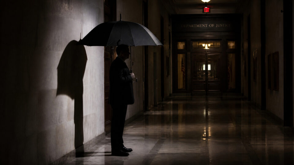 A man in a suit holding an umbrella stands in a dimly lit courthouse hallway, casting a shadow on the wall, with the Justice Department sign visible in the background.