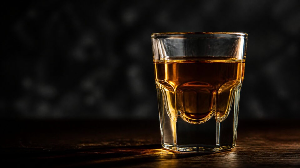 How to Choose Shot Glasses: Why a 2-Oz Glass Holds Only 1.5 2 A clear glass filled with amber whiskey placed on a dark wooden surface with a blurred dark background.
